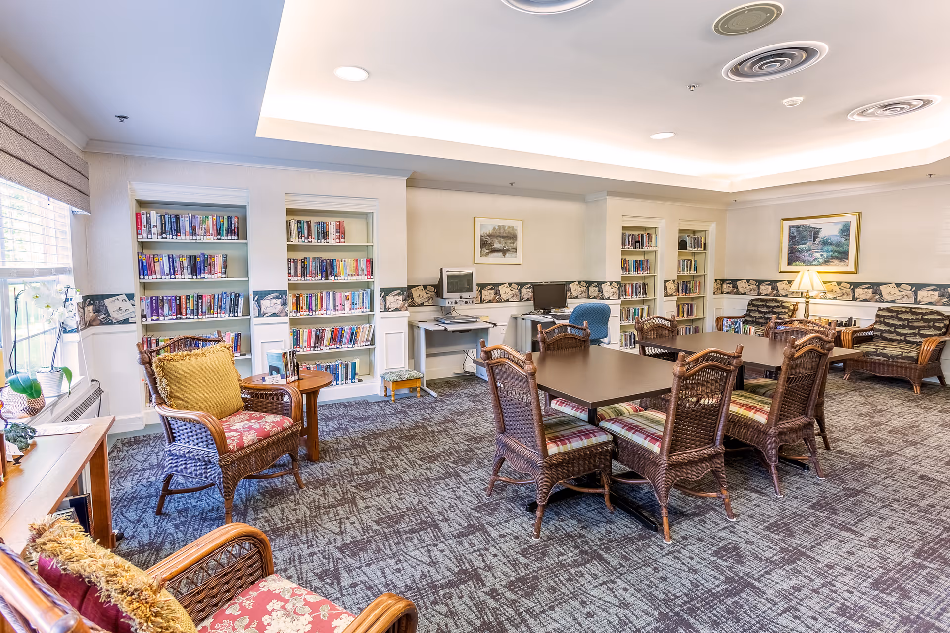 A bright and cozy library or common room with bookshelves filled with books along the walls, wicker chairs with cushions, two tables with wicker chairs around them, a computer desk with two computers, and a window letting in natural light.