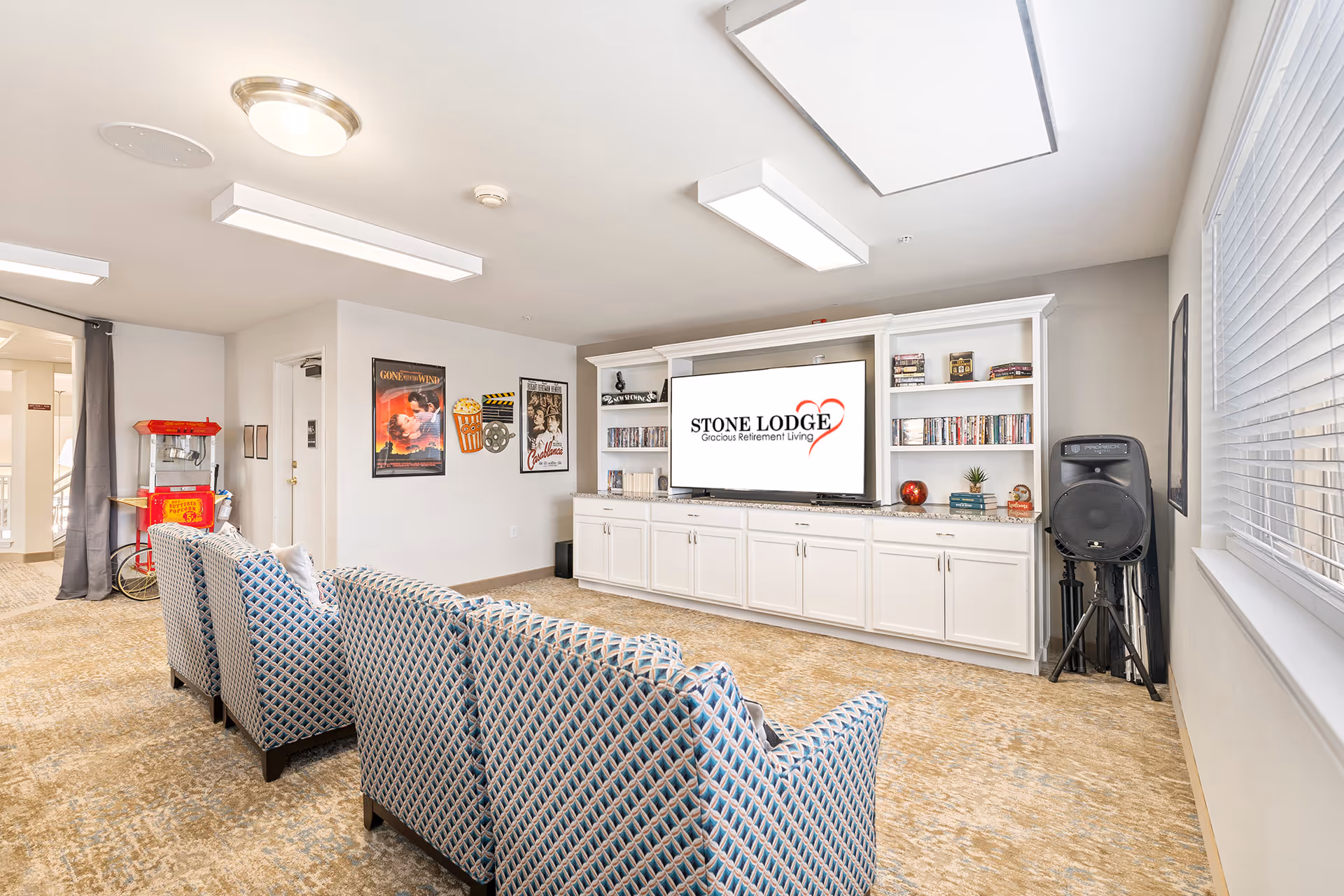 A cozy entertainment room with patterned armchairs facing a large flat-screen TV mounted on a white built-in cabinet. The TV screen displays the logo for Stone Lodge Gracious Retirement Living. The room has movie posters on the wall, a popcorn machine in the corner, shelves with DVDs and decorative items, a large speaker, and a window with blinds letting in natural light.