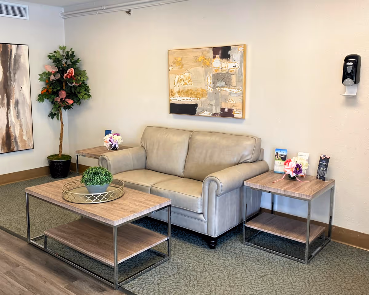 A cozy sitting area with a light gray leather loveseat, a wooden coffee table with a decorative tray and small plant, and a matching wooden side table with brochures and a small flower arrangement. There is a tall artificial plant in a black pot in the corner and two abstract paintings hanging on the light-colored walls. A wall-mounted hand sanitizer dispenser is visible on the right side.