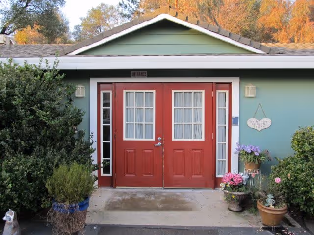 Front entrance of a single-story building with double red doors, sidelights, green siding, and potted plants.