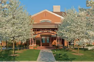Front exterior view of a senior living community building with a covered entrance, surrounded by green grass and blooming trees on either side of a concrete walkway.