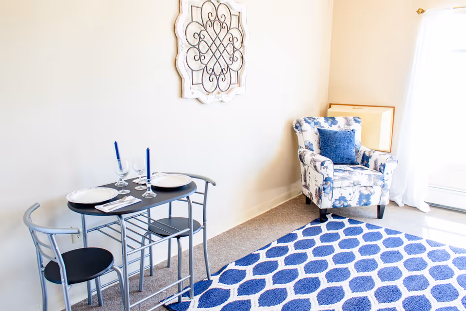 A small dining area with a black metal table set for two with plates, napkins, wine glasses, and blue candles. Next to it is a floral patterned armchair with a blue cushion, placed on a blue and white patterned rug. The room has light-colored walls and a decorative wall hanging above the table.