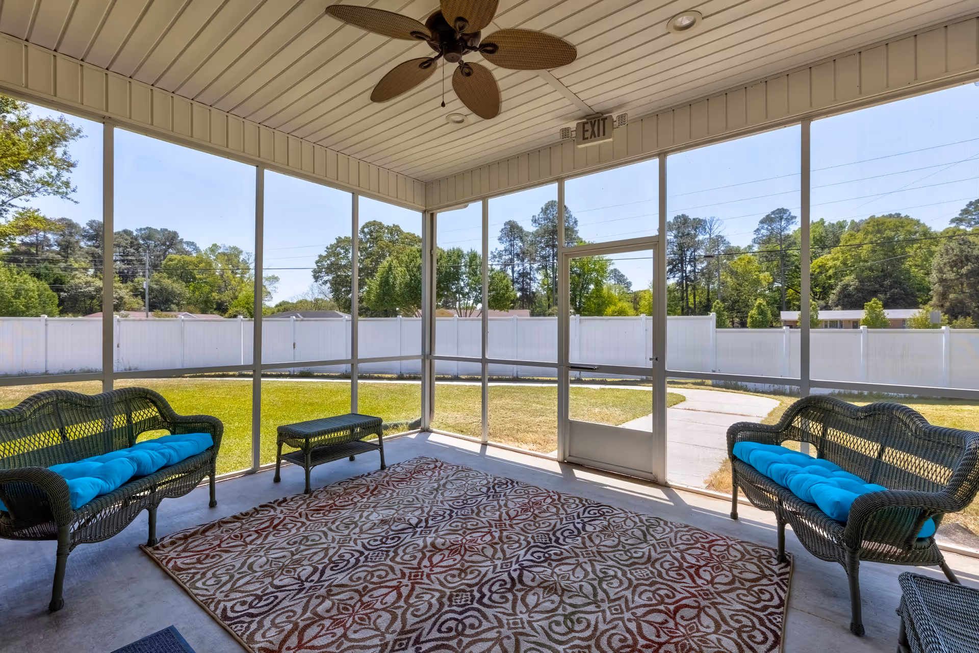 A screened-in porch area with two wicker benches with blue cushions, a wicker ottoman, a patterned area rug, and a ceiling fan. The porch overlooks a grassy yard enclosed by a white fence with trees in the background.