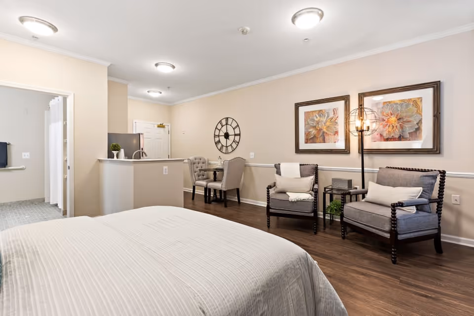 A cozy senior living facility room featuring a bed with a light-colored bedspread in the foreground, two cushioned armchairs with pillows and a small side table with a lamp and decorative items against the wall, two framed floral artworks above the chairs, a small dining table with two upholstered chairs near the back wall, and a kitchenette area with a sink and refrigerator. The room has wooden flooring and neutral-colored walls. A bathroom with a shower curtain is visible through an open door on the left.