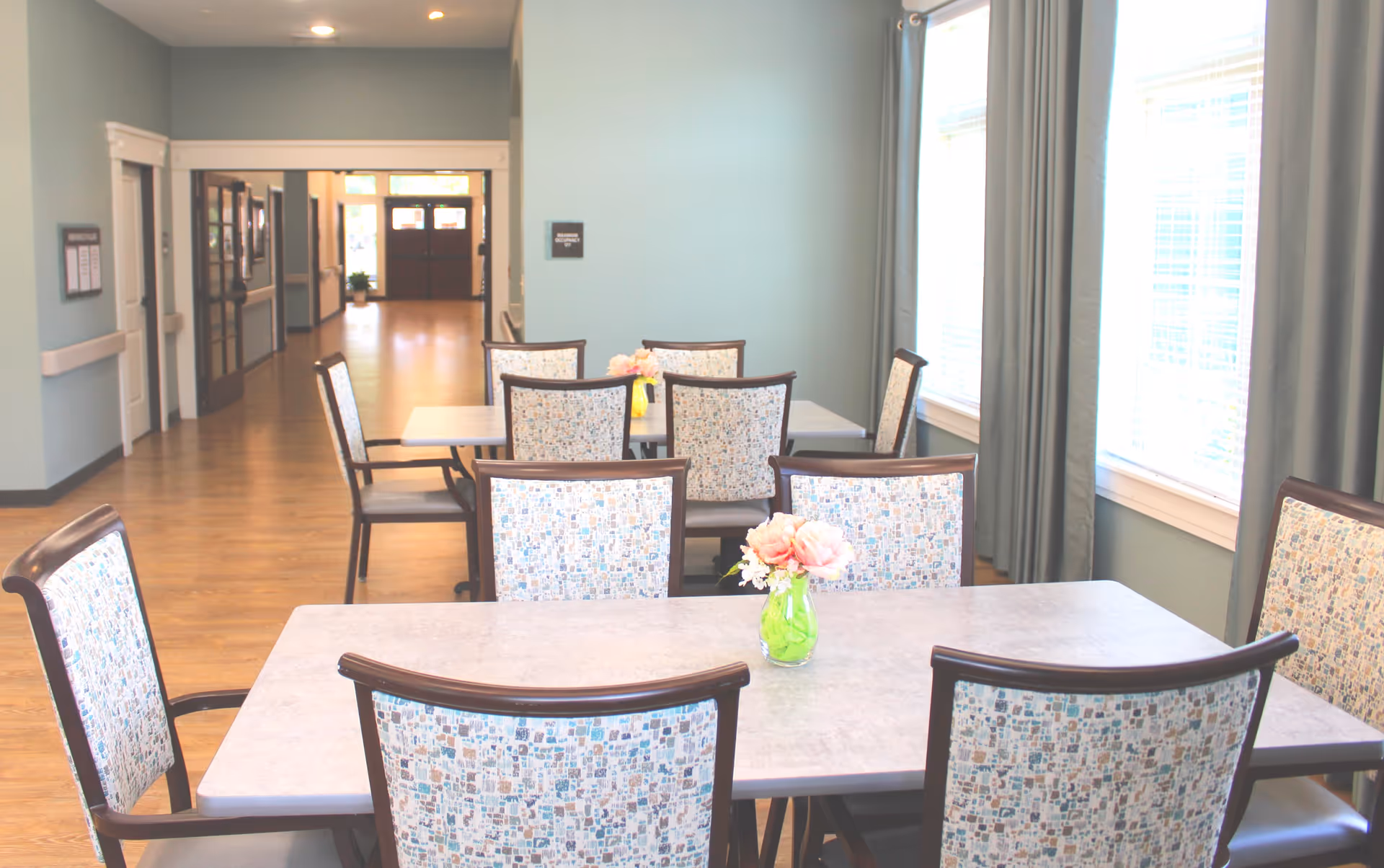 A bright dining area with two rectangular tables, each surrounded by six chairs with patterned upholstery. A small green vase with pink flowers is placed on the nearest table. Large windows with curtains allow natural light to fill the room. The background shows a hallway with wooden flooring and doors.