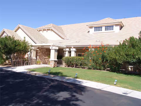 Exterior view of a single-story building with a beige tiled roof, stone and stucco walls, surrounded by green bushes and a small lawn under a clear blue sky.