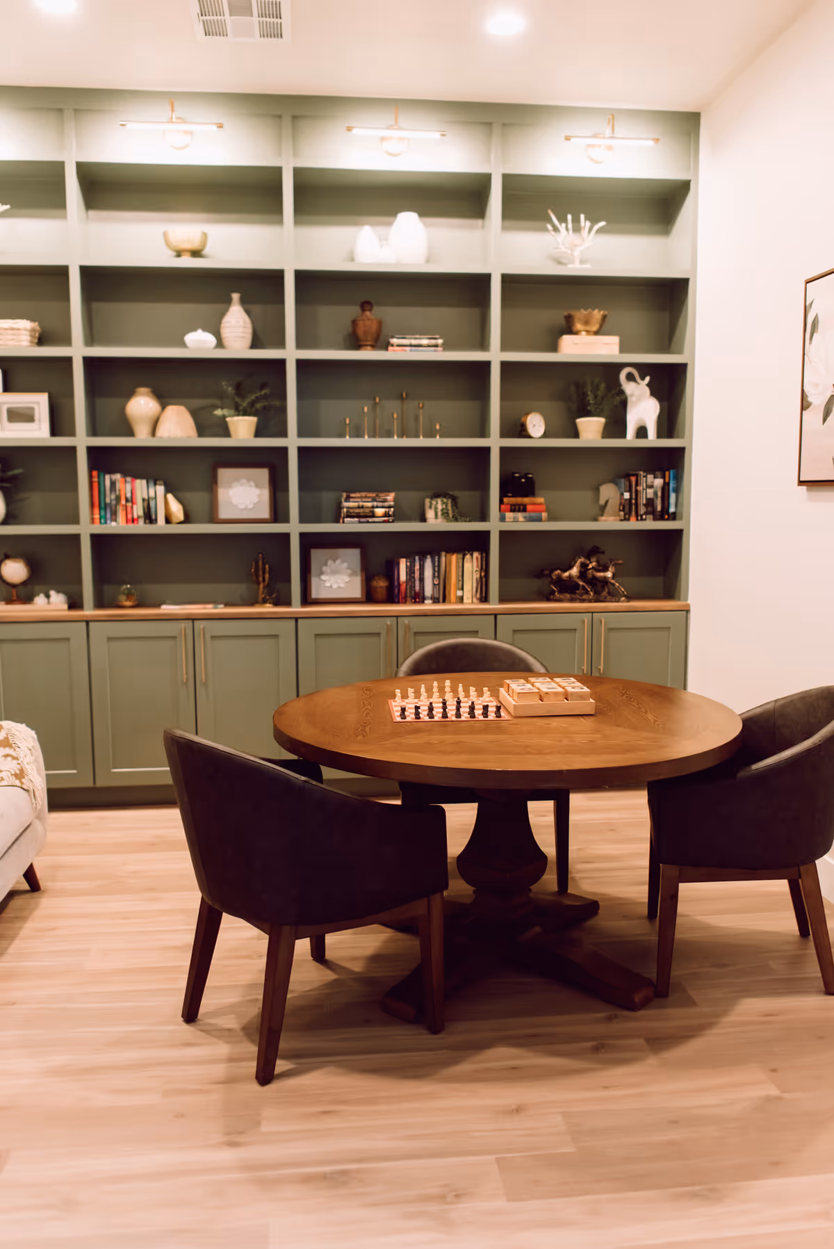 A cozy room with a round wooden table in the center surrounded by three dark upholstered chairs. On the table, there is a chessboard and a wooden box. Behind the table, there is a large built-in shelving unit painted green, filled with various decorative items, books, and plants. The room has light wood flooring and soft lighting from ceiling fixtures.