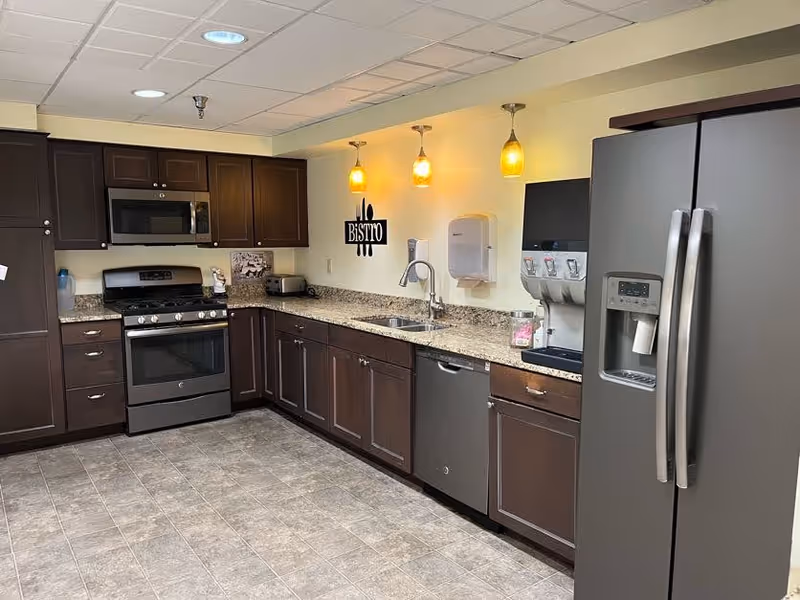 A modern kitchen with dark wood cabinets, granite countertops, and stainless steel appliances including a refrigerator, dishwasher, microwave, and stove. Three pendant lights hang above the sink area, and a wall decoration reads 'Bistro'.