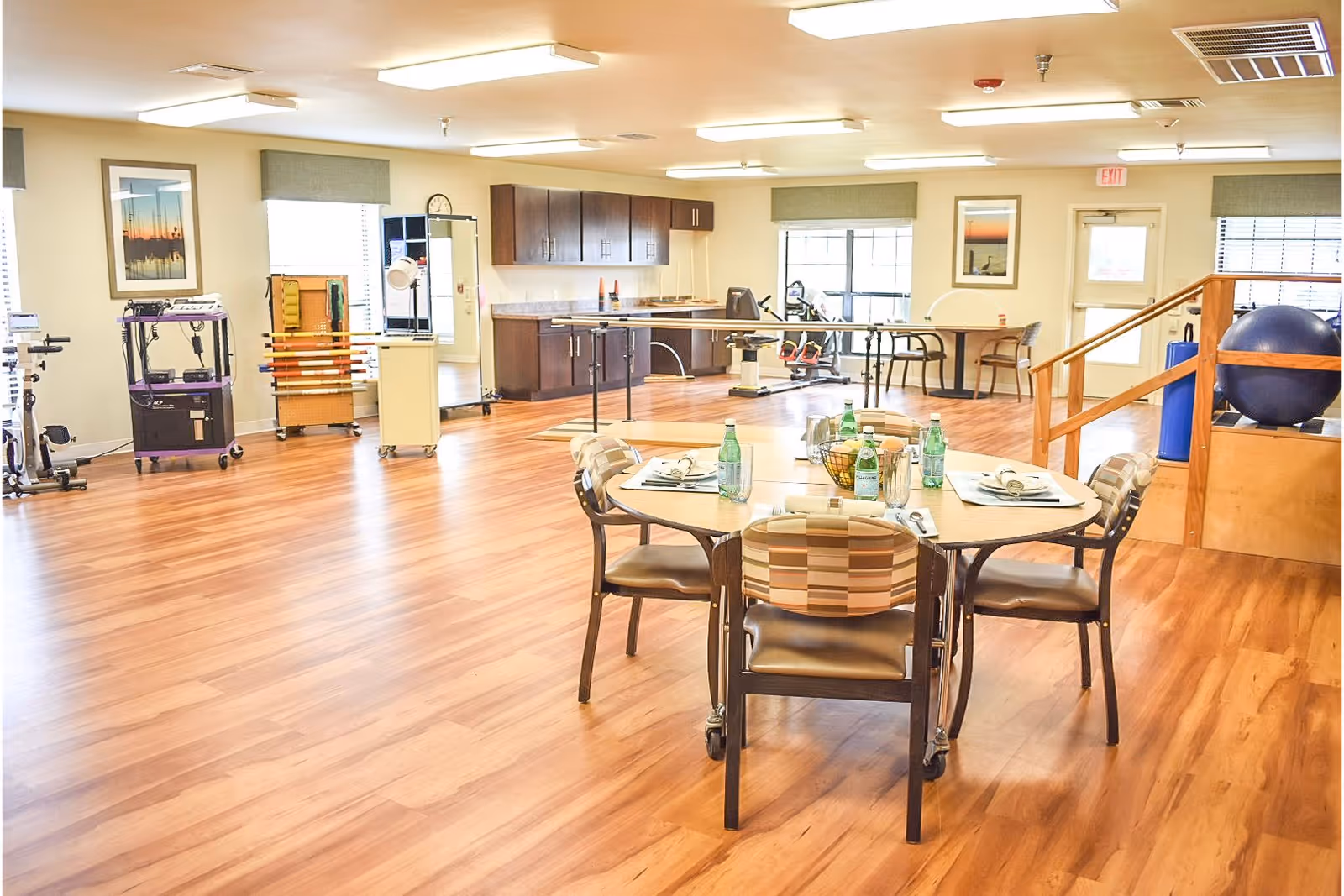 A spacious room with wooden flooring featuring a table set for four with water bottles and napkins. The room has exercise equipment, cabinets, and large windows with green valances. There is a staircase with a wooden railing and a large exercise ball on the right side.