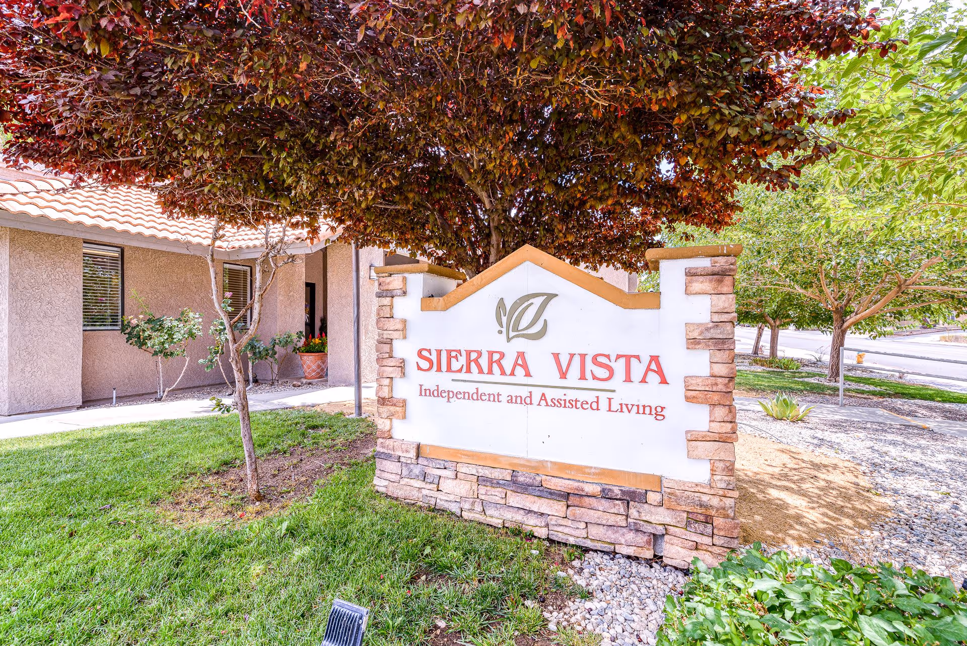Outdoor view of the entrance area of Sierra Vista Independent and Assisted Living facility, featuring a stone and stucco sign with the facility name, surrounded by green grass, trees, and landscaping.
