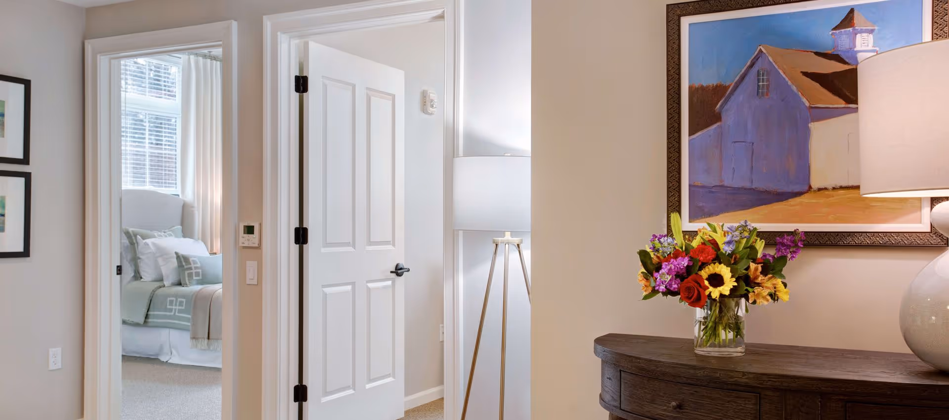 Hallway interior with an open door revealing a bedroom and a console table topped with a lamp and vase of flowers.