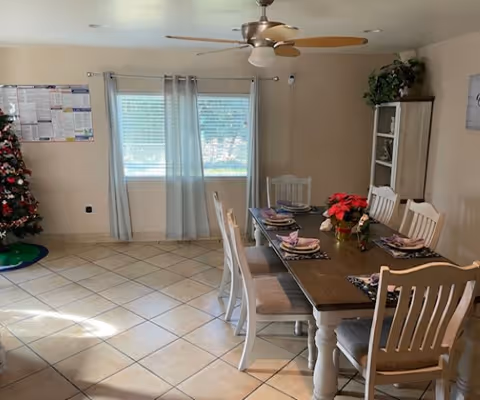 Bright dining room with a wooden table set for six, poinsettia centerpiece, ceiling fan, window with light curtains and a Christmas tree in the corner.