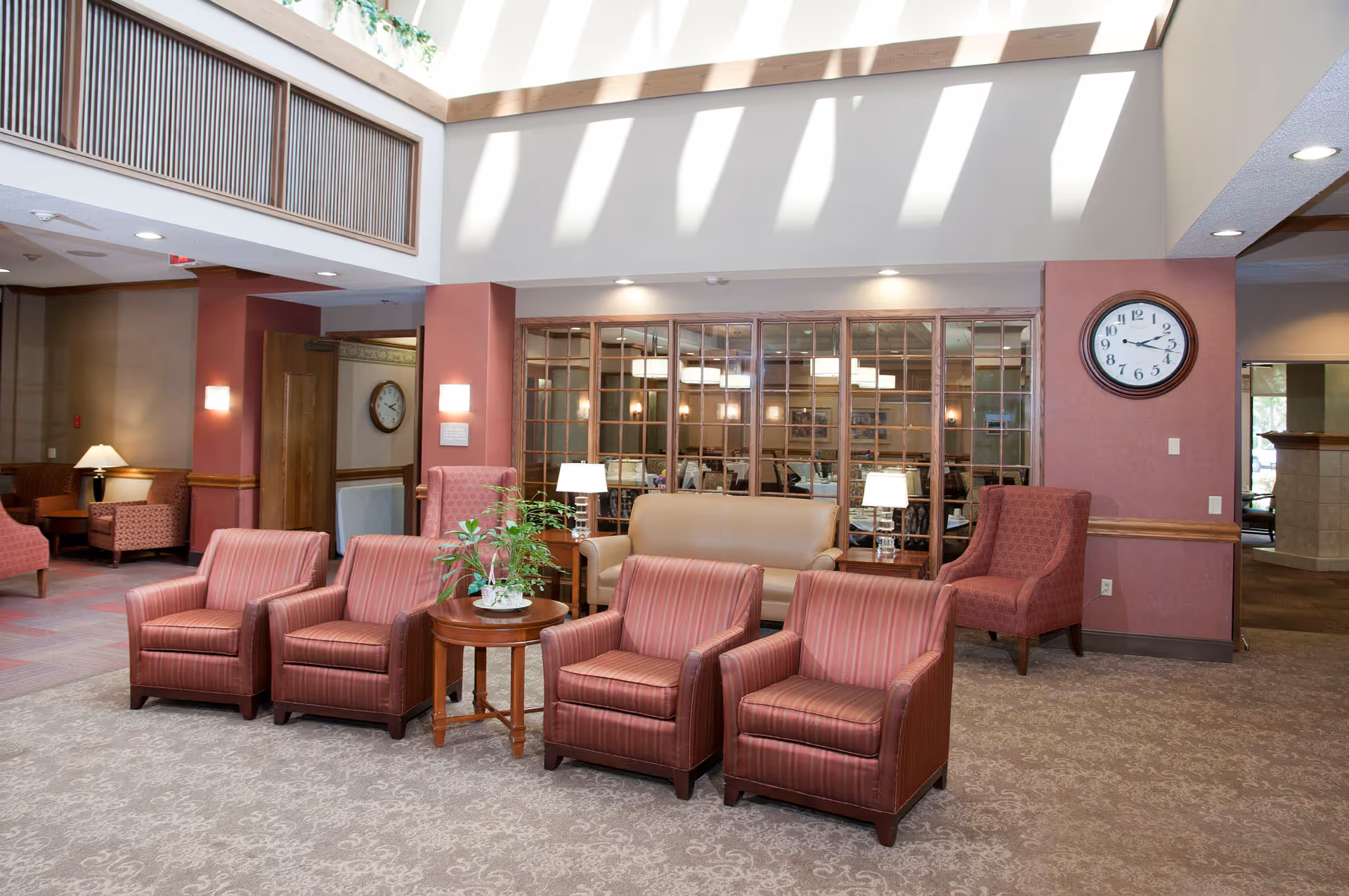 A spacious senior living facility lounge area with several upholstered armchairs and a sofa arranged around small wooden tables. The room has a high ceiling with skylights casting shadows on the walls. There is a large clock on the wall, lamps on side tables, and a glass partition separating this area from a dining room visible in the background.