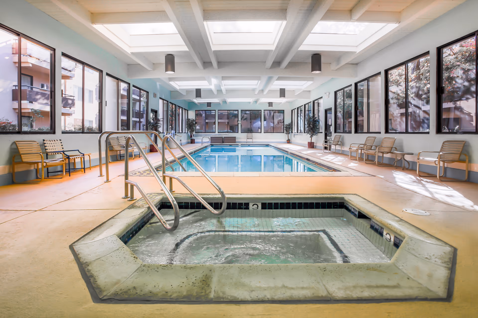 Indoor swimming pool area with a hot tub in the foreground. The room has large windows on both sides, allowing natural light to fill the space. There are several chairs and small tables lined up along the walls. The ceiling features multiple skylights and hanging light fixtures.