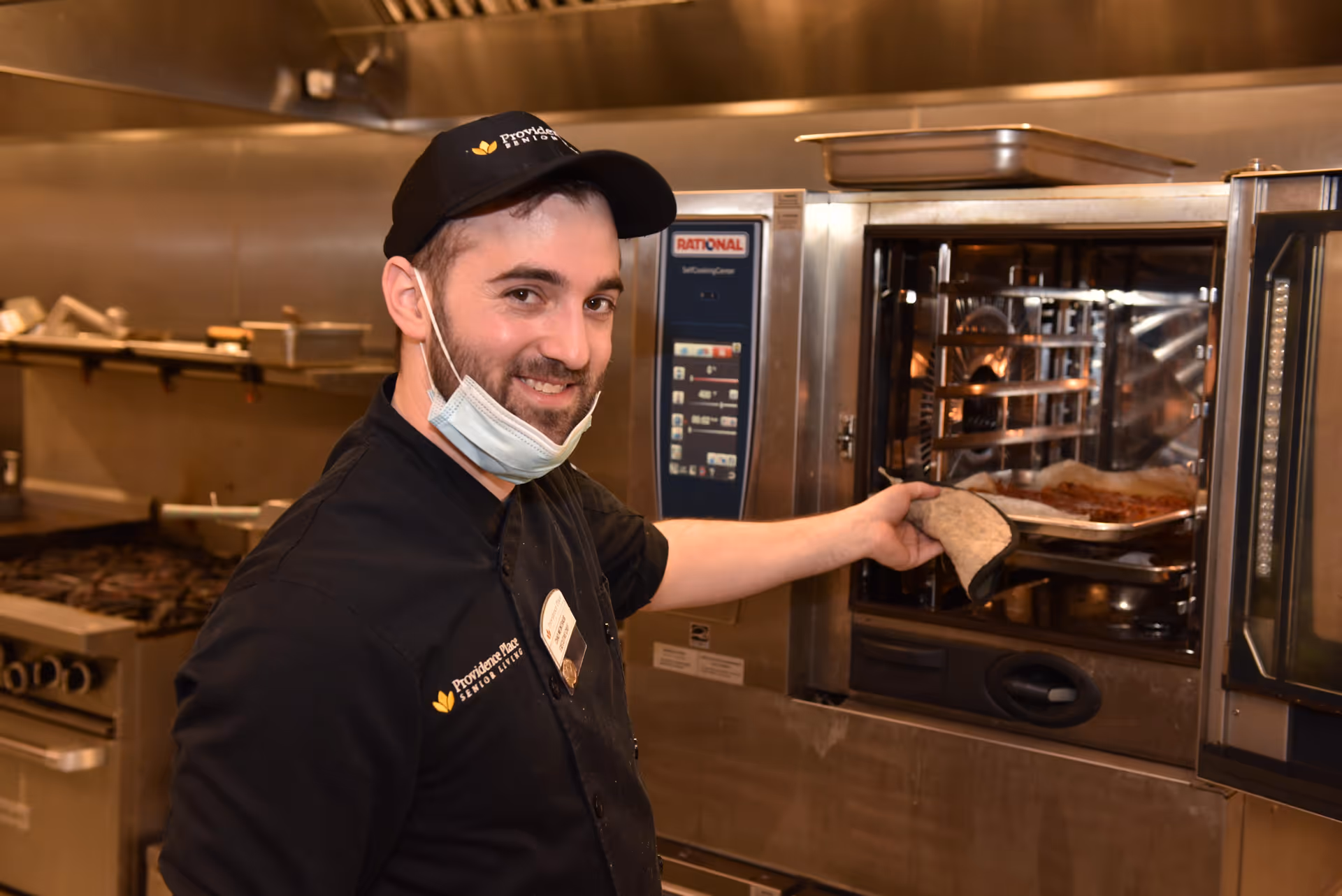 A smiling cook wearing a Providence Place cap and lowered face mask places a tray into a commercial oven in a professional kitchen.