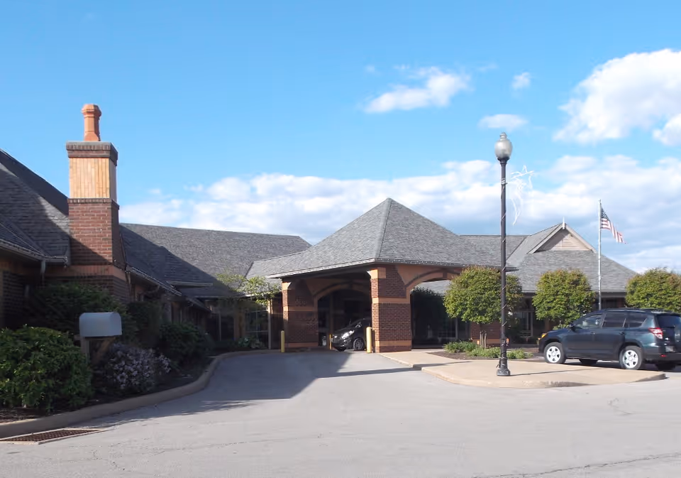 Exterior view of Orchard Manor facility showing a brick building with a covered entrance, a driveway, parked cars, a lamppost, and an American flag on a flagpole under a partly cloudy blue sky.