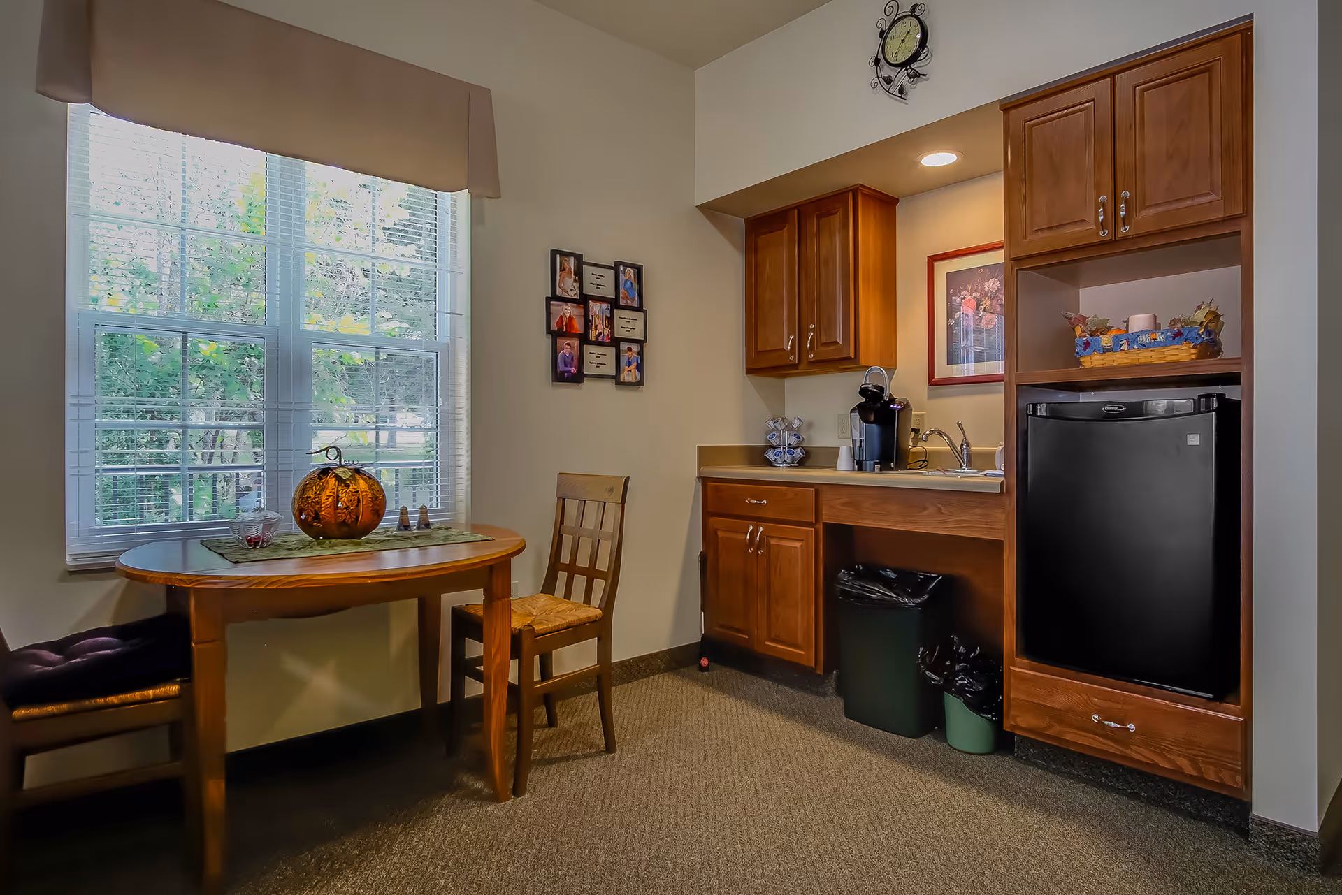 A small kitchenette area with wooden cabinets, a black mini refrigerator, a coffee maker, and a sink. Next to the kitchenette is a wooden table with two chairs, one with a cushion. A window with blinds and a valance lets in natural light, and a decorative pumpkin sits on the table. A wall clock and framed pictures are visible on the walls.