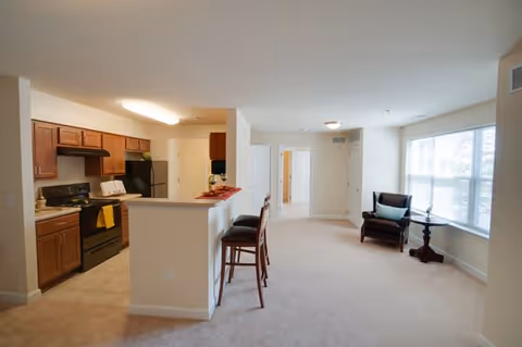Open-plan apartment interior showing a kitchen with a breakfast bar and stools and a carpeted living area with chairs by a large window.