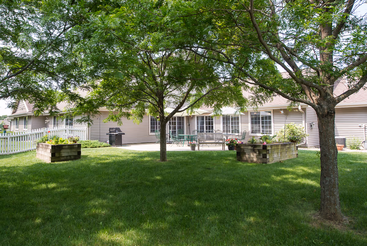 Outdoor view of a senior living facility with green grass, two trees, raised garden beds with flowers, and a patio area with tables, chairs, and a bench in front of a beige building with multiple windows.