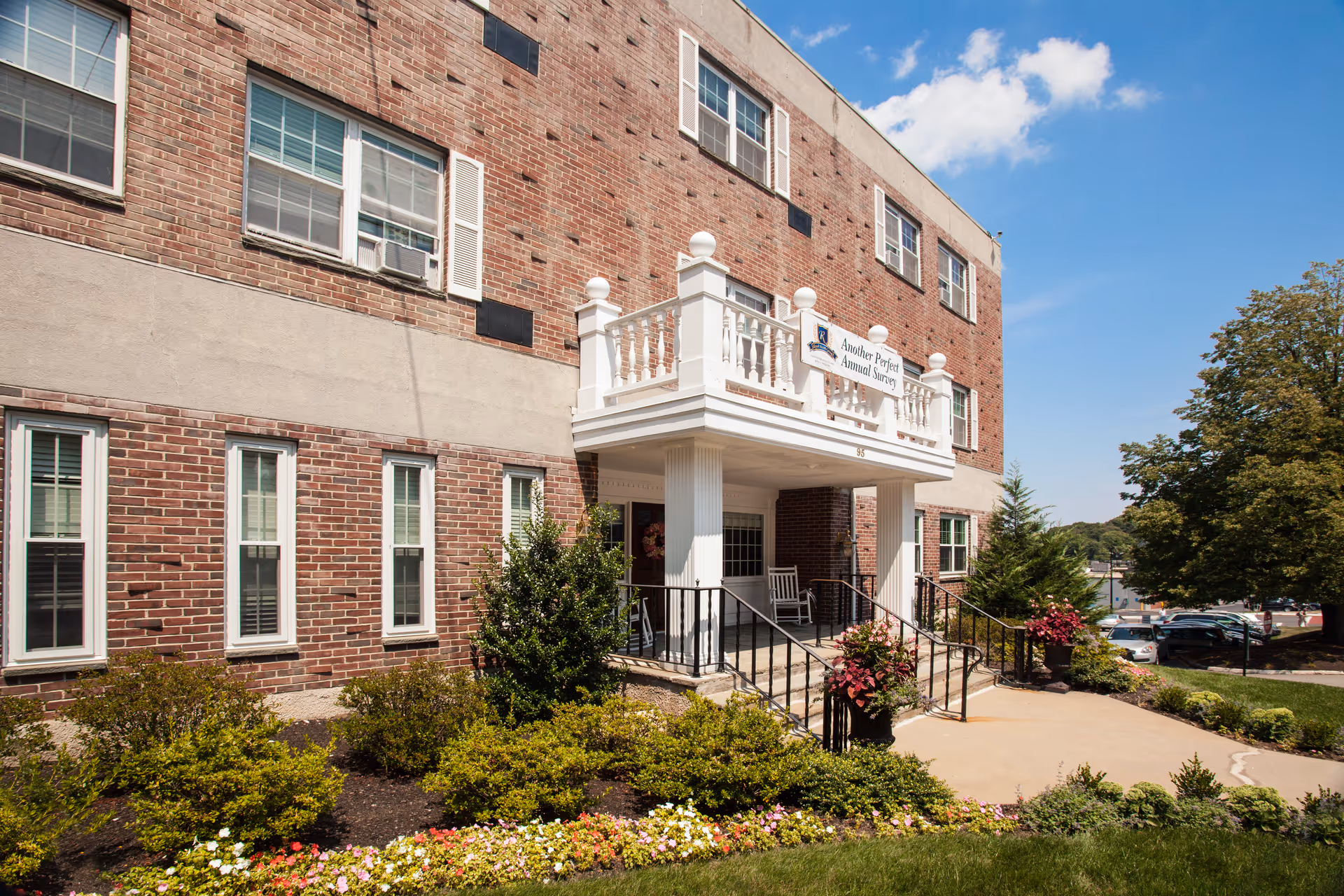 Exterior view of a brick building with a white balcony and entrance porch. There are stairs with black railings leading up to the entrance, surrounded by well-maintained bushes and colorful flowers. The sky is clear with a few clouds, and there are trees and parked cars visible in the background.