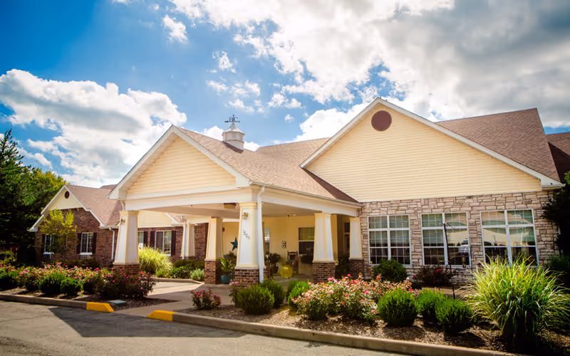Exterior view of a single-story senior living facility building with a covered entrance, beige siding, stone accents, and a landscaped garden with bushes and flowers under a partly cloudy sky.