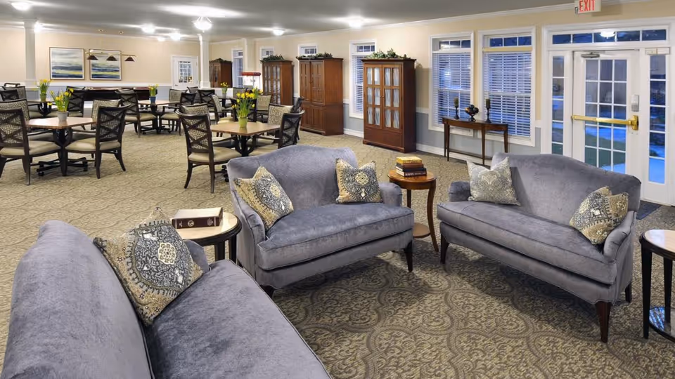 A spacious senior living common area with three gray upholstered sofas arranged around small wooden tables with books and decorative pillows. In the background, there are multiple dining tables with chairs and vases of yellow flowers. The room has beige walls, large windows with white blinds, and carpeted floors with a patterned design. Wooden cabinets and framed artwork are visible along the walls.