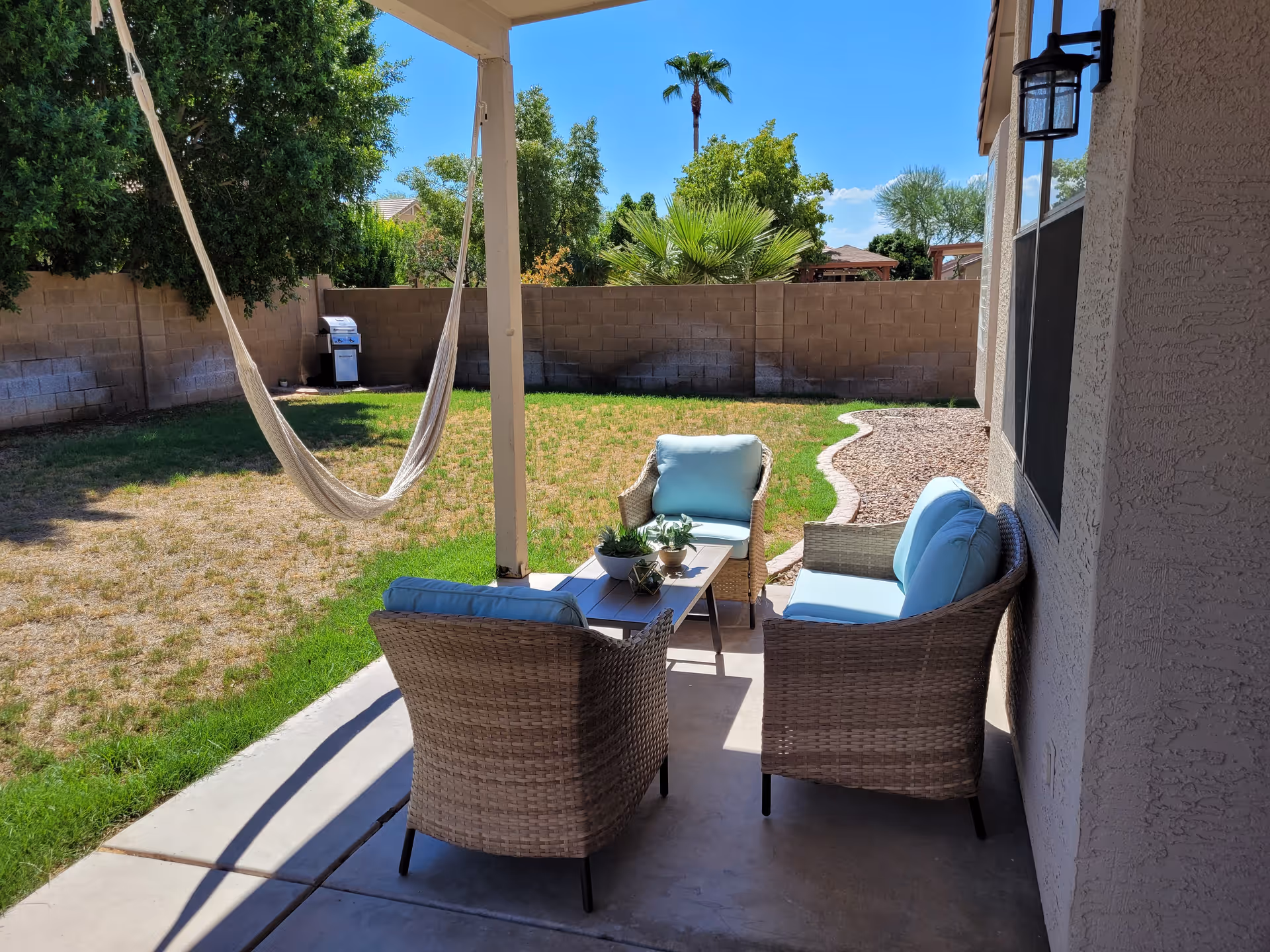 Backyard patio with wicker seating and blue cushions under a covered porch, a hammock, lawn, and a grill.