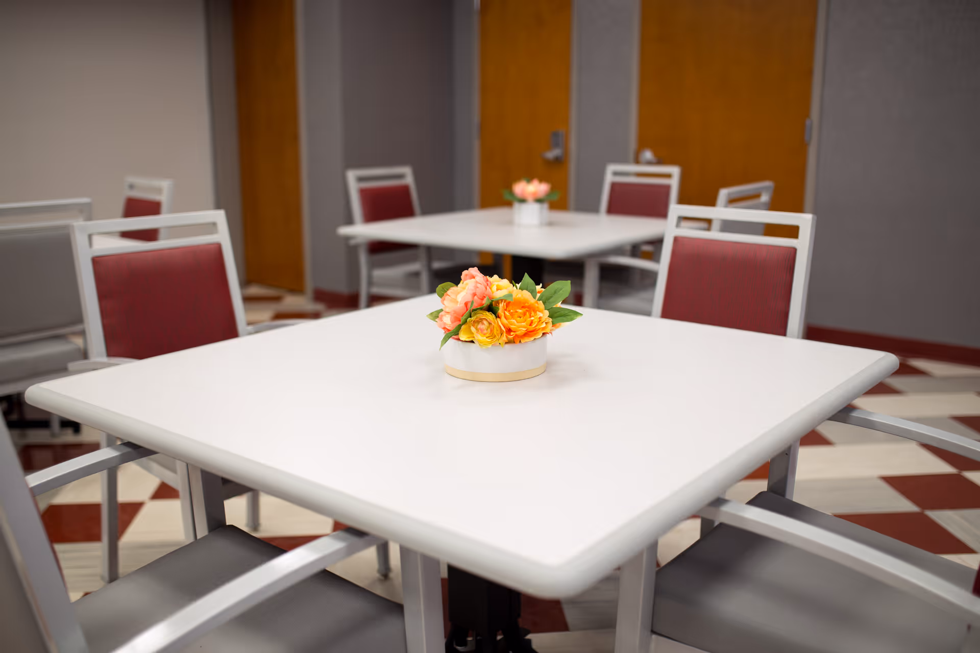Square dining table with a small floral centerpiece and surrounding chairs in a communal dining room.