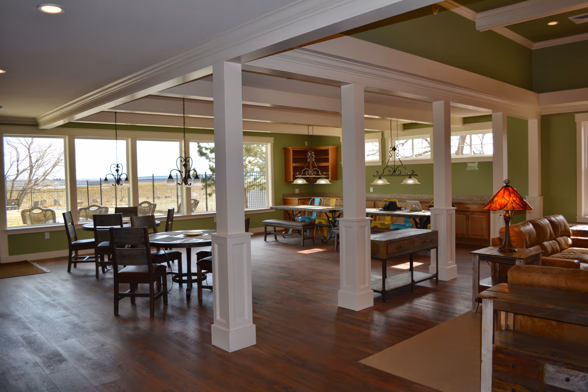 Interior view of a spacious room with green walls and large windows letting in natural light. The room features a dining area with wooden tables and chairs, a seating area with brown leather sofas, and hanging light fixtures. There are white pillars and wooden flooring throughout the space.