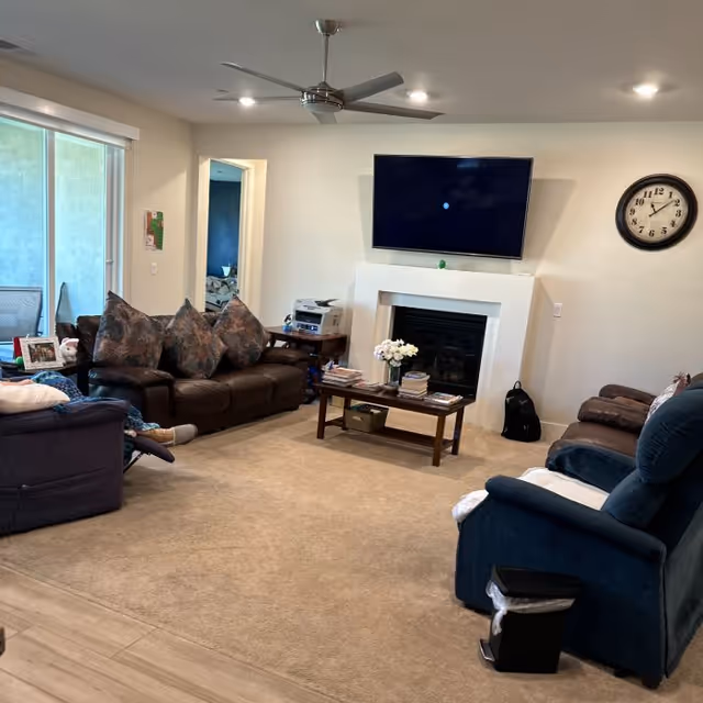 A cozy living room with a brown leather sofa, a blue recliner, and another cushioned chair arranged around a wooden coffee table with books and a vase of flowers. A flat-screen TV is mounted above a white fireplace on the wall. A round clock is hung on the wall to the right. There is a ceiling fan with lights above, and a sliding glass door on the left side of the room.