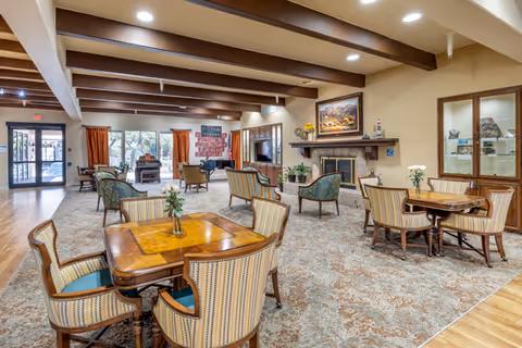 Spacious senior living common room with card tables and upholstered chairs, a fireplace, exposed wooden ceiling beams, and large windows.
