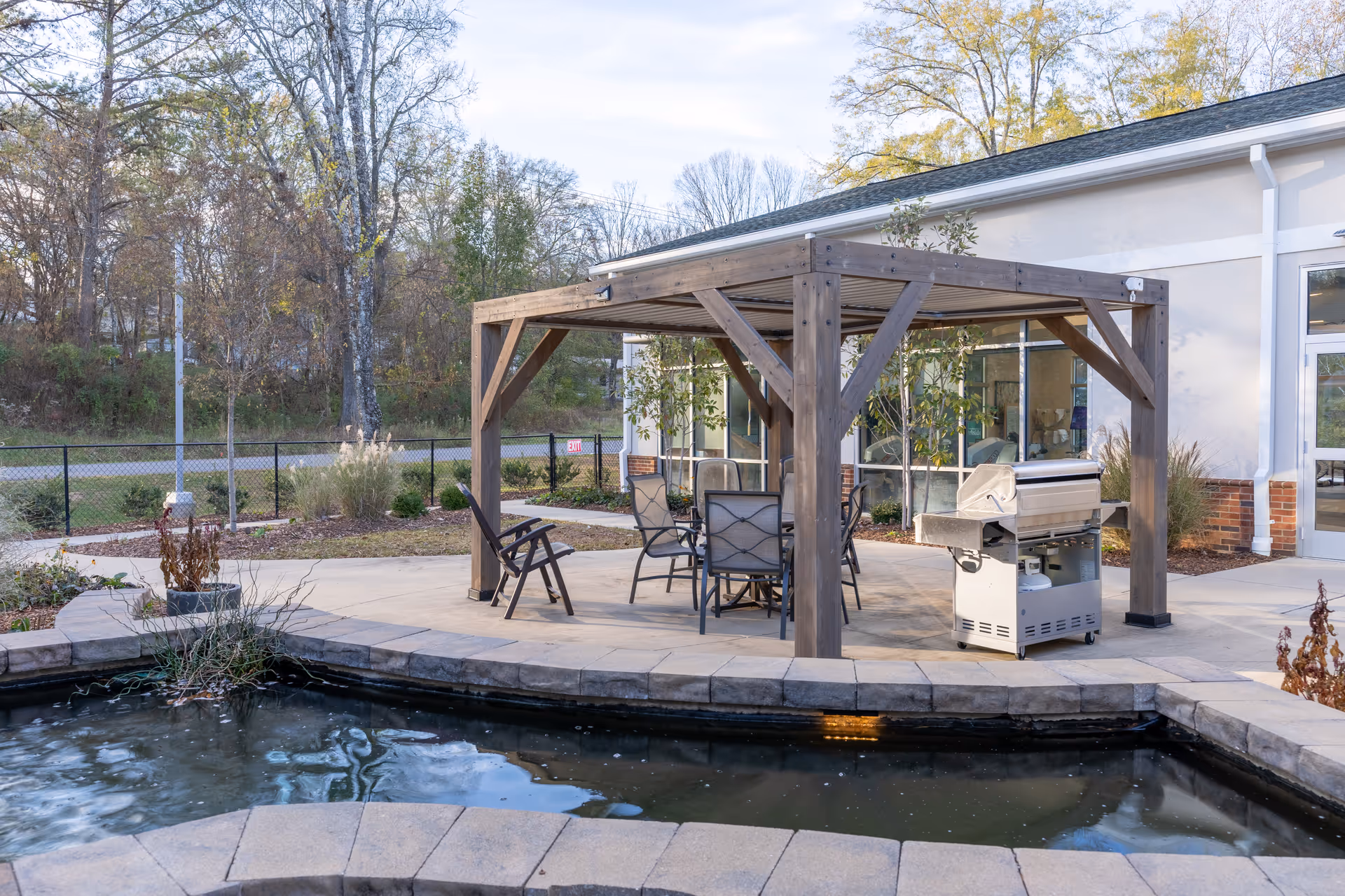 Outdoor patio area with a wooden pergola covering a table and chairs, a grill nearby, and a small pond with a stone border in the foreground. Trees and a building with large windows are visible in the background.