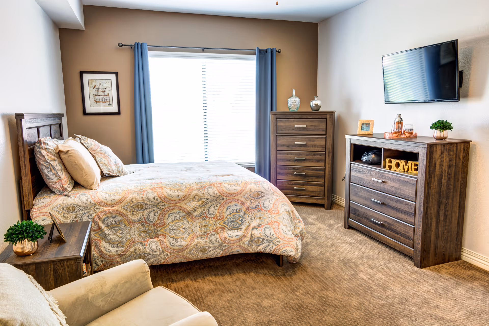 A cozy bedroom with a large window covered by blue curtains. The room features a bed with a patterned bedspread and multiple pillows, a wooden nightstand with a small plant, a wooden dresser, a wooden chest of drawers, a wall-mounted flat-screen TV, and a beige armchair in the foreground.
