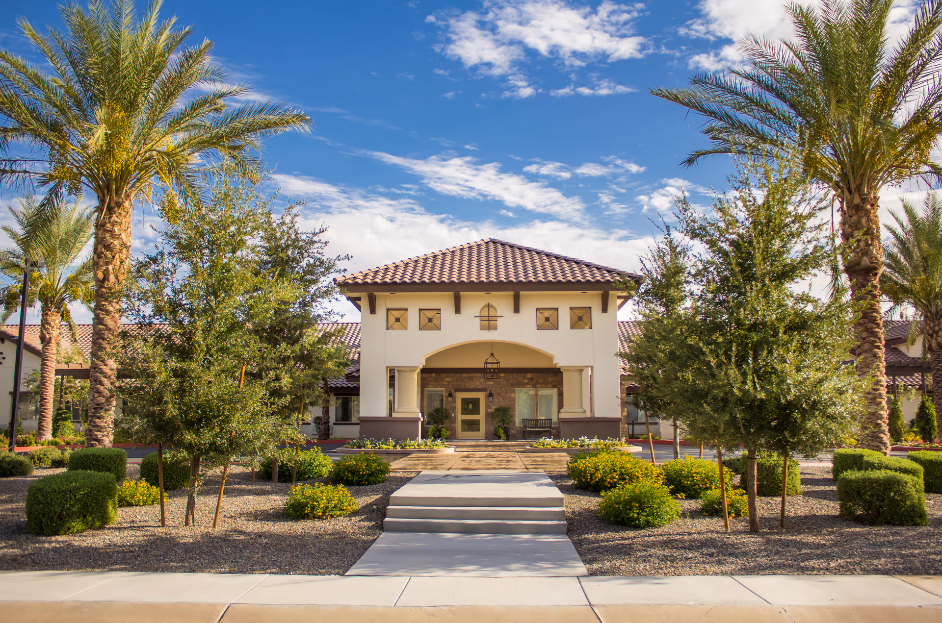 Front exterior view of Amber Creek Inn Memory Care Community building with a tiled roof, surrounded by palm trees, bushes, and landscaped garden under a partly cloudy blue sky.