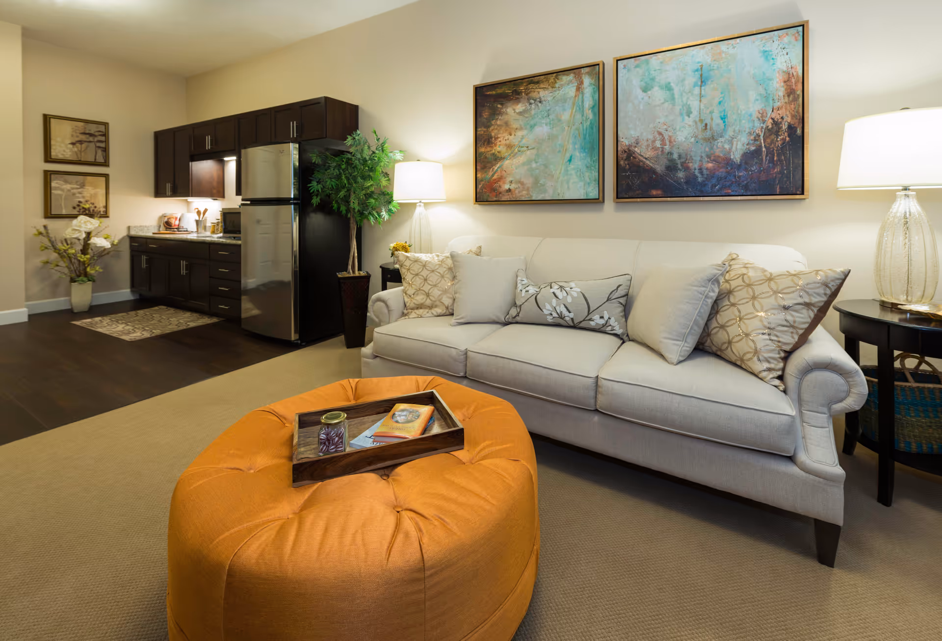 A cozy living room area with a beige sofa adorned with decorative pillows, two abstract paintings on the wall above, and a glass table lamp on a side table. In front of the sofa is a large orange tufted ottoman with a wooden tray holding a small jar and a book. To the left, there is a compact kitchen area with dark wood cabinets, a stainless steel refrigerator, and a countertop with kitchen utensils. The room has warm lighting and a beige carpet.