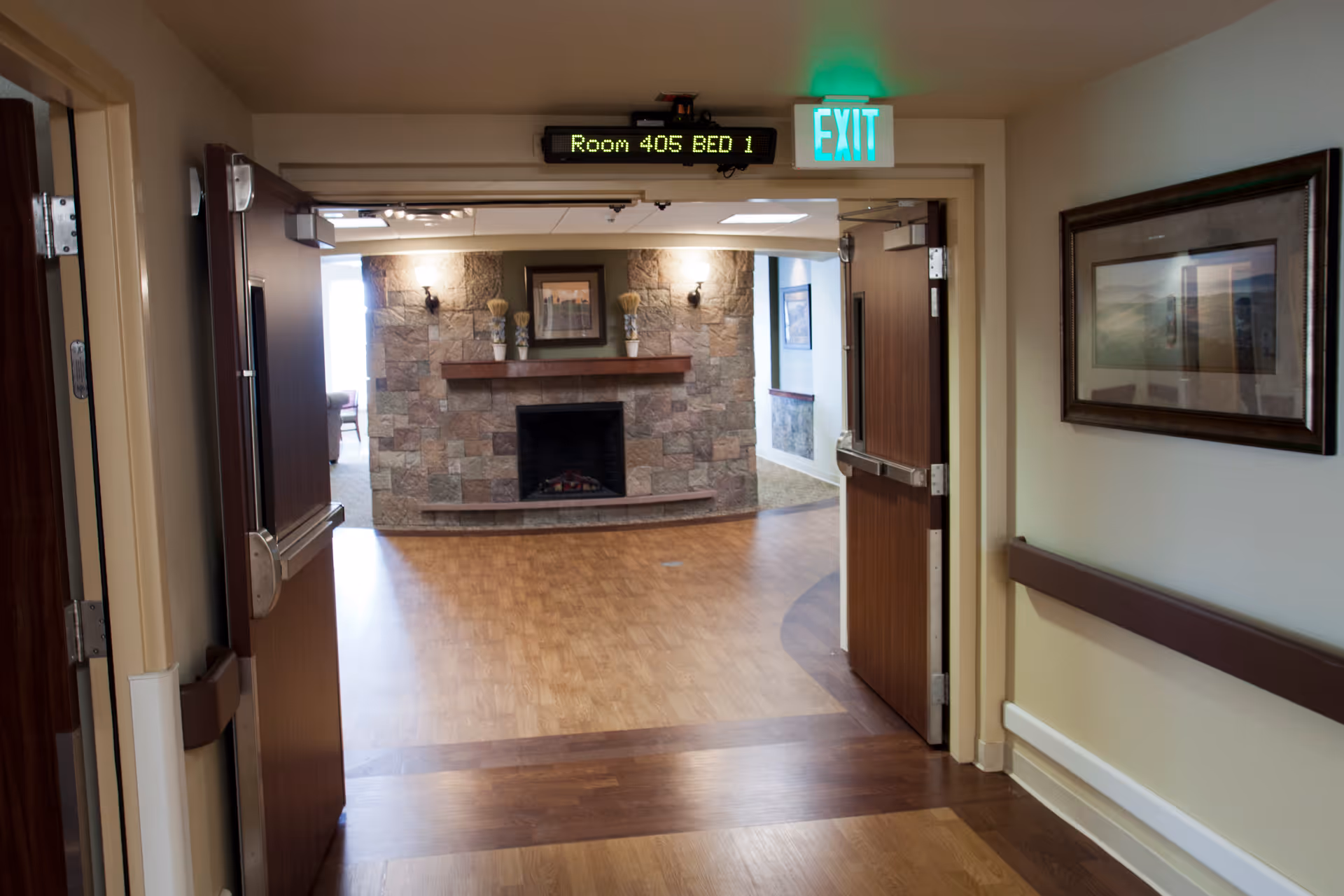 View through double doors into a room with a stone fireplace, wooden mantel, and framed artwork above it. The floor is wood, and there is an exit sign and a digital sign above the doors reading 'Room 405 BED 1'. A framed picture hangs on the right wall of the hallway leading to the room.