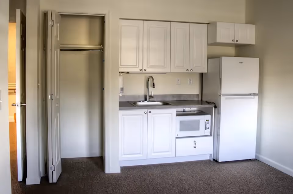 Small kitchenette area with white cabinets, a sink, a microwave, and a refrigerator. To the left of the kitchenette is an open closet with bi-fold doors. The floor is carpeted and the walls are painted light beige.