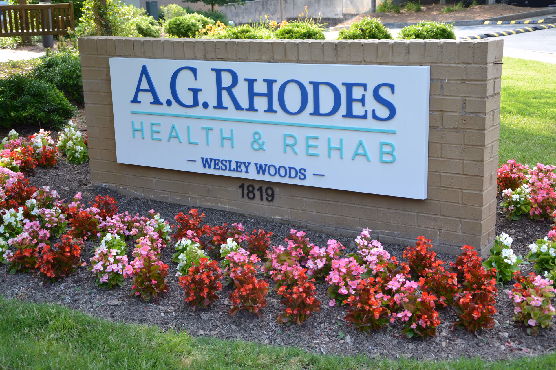 A brick sign for A.G. Rhodes Health & Rehab Wesley Woods surrounded by a flower bed with red, pink, and white flowers and green grass.