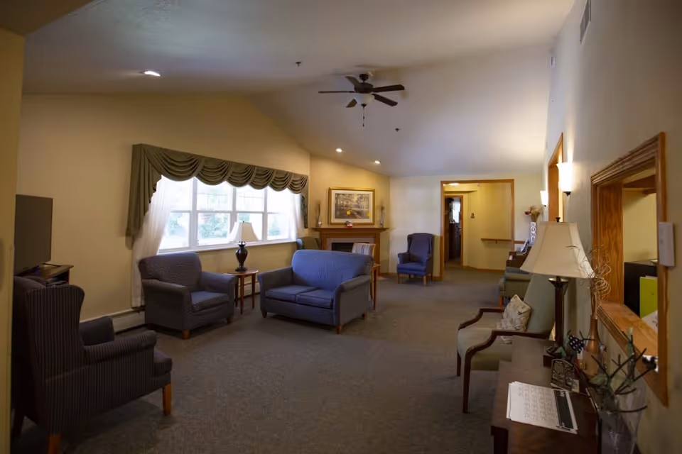 A spacious living room area in an assisted living facility with multiple upholstered chairs and a loveseat arranged around a window with green valance curtains. There is a ceiling fan, a fireplace with a framed picture above it, and a TV on a stand. A table with a lamp and decorative items is visible on the right side of the room.