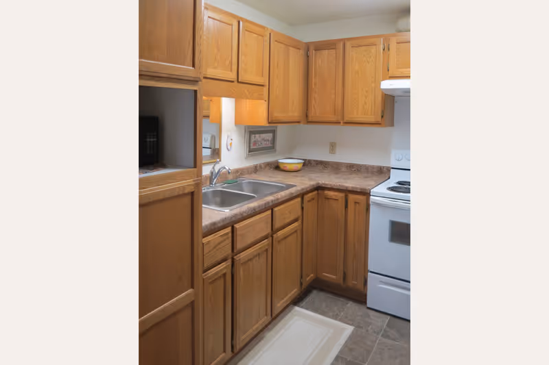 A compact kitchen with wooden cabinets, a double stainless steel sink, a white electric stove with oven, and a countertop with a decorative bowl. The floor has a small rug, and there is a framed picture on the wall above the counter.
