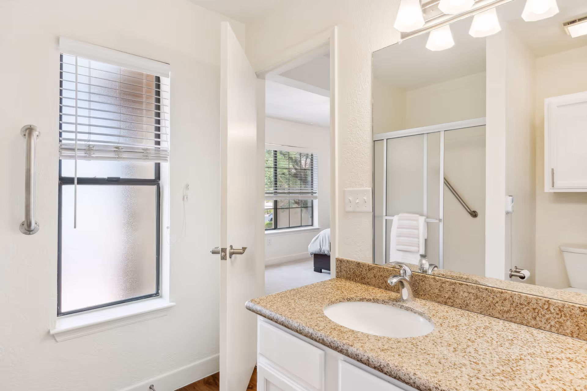 Bright bathroom with a granite countertop sink, large mirror, frosted window with blinds, and a partially open door revealing a bedroom with a bed and window. The bathroom also features a shower with sliding glass doors and a grab bar.