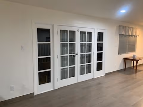 Empty interior room featuring white French doors with glass panes, wood-look flooring, and a small table beneath a window.
