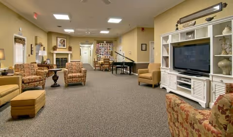 A spacious living room area in a senior living facility with multiple upholstered chairs and ottomans arranged around a carpeted floor. There is a large white entertainment center with a flat-screen TV on the right side, a fireplace with framed artwork above it on the left, and a black grand piano in the background. The walls are painted a warm beige color, and the room is well-lit with ceiling lights.