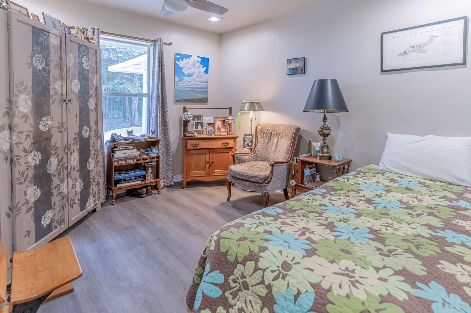 A cozy bedroom in an assisted living facility featuring a bed with a floral patterned quilt, a cushioned armchair, a wooden side table with a lamp, a small wooden cabinet with framed photos, a bookshelf with books, a large window with curtains, and a wardrobe with floral designs.