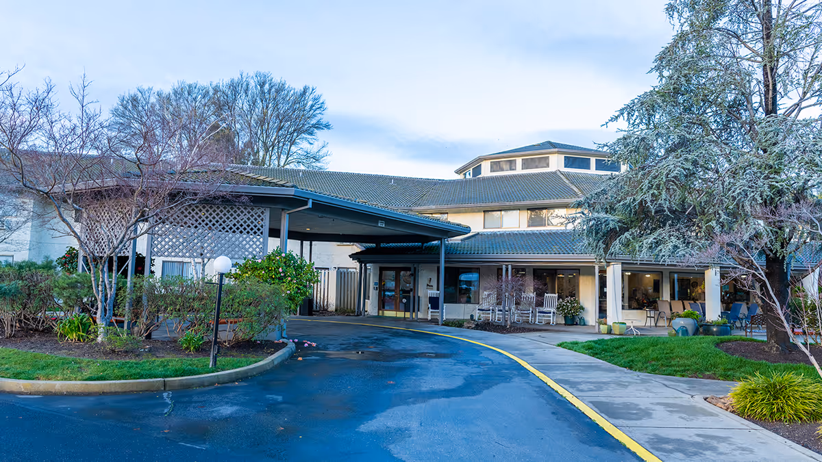 Exterior view of Provincial Chico Senior Living facility showing the entrance with a covered drop-off area, surrounded by landscaped bushes, trees, and a paved driveway with a yellow curb.