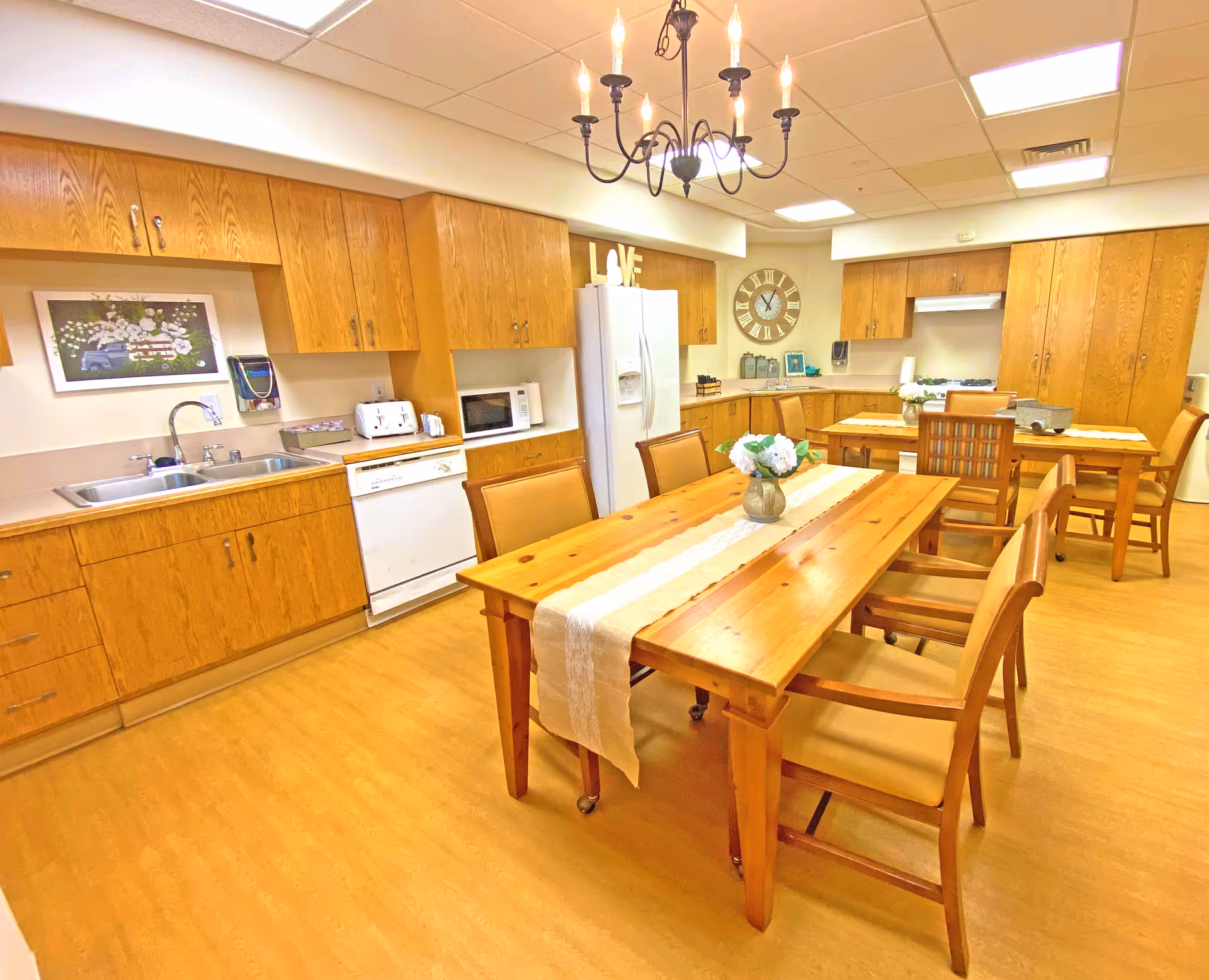 Communal kitchen and dining area with wooden cabinets, a long wooden table and chairs, appliances, and an overhead chandelier.