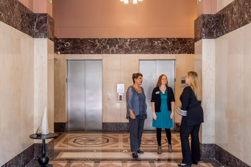Three women talking in a marble-floored elevator lobby with closed stainless steel elevator doors.