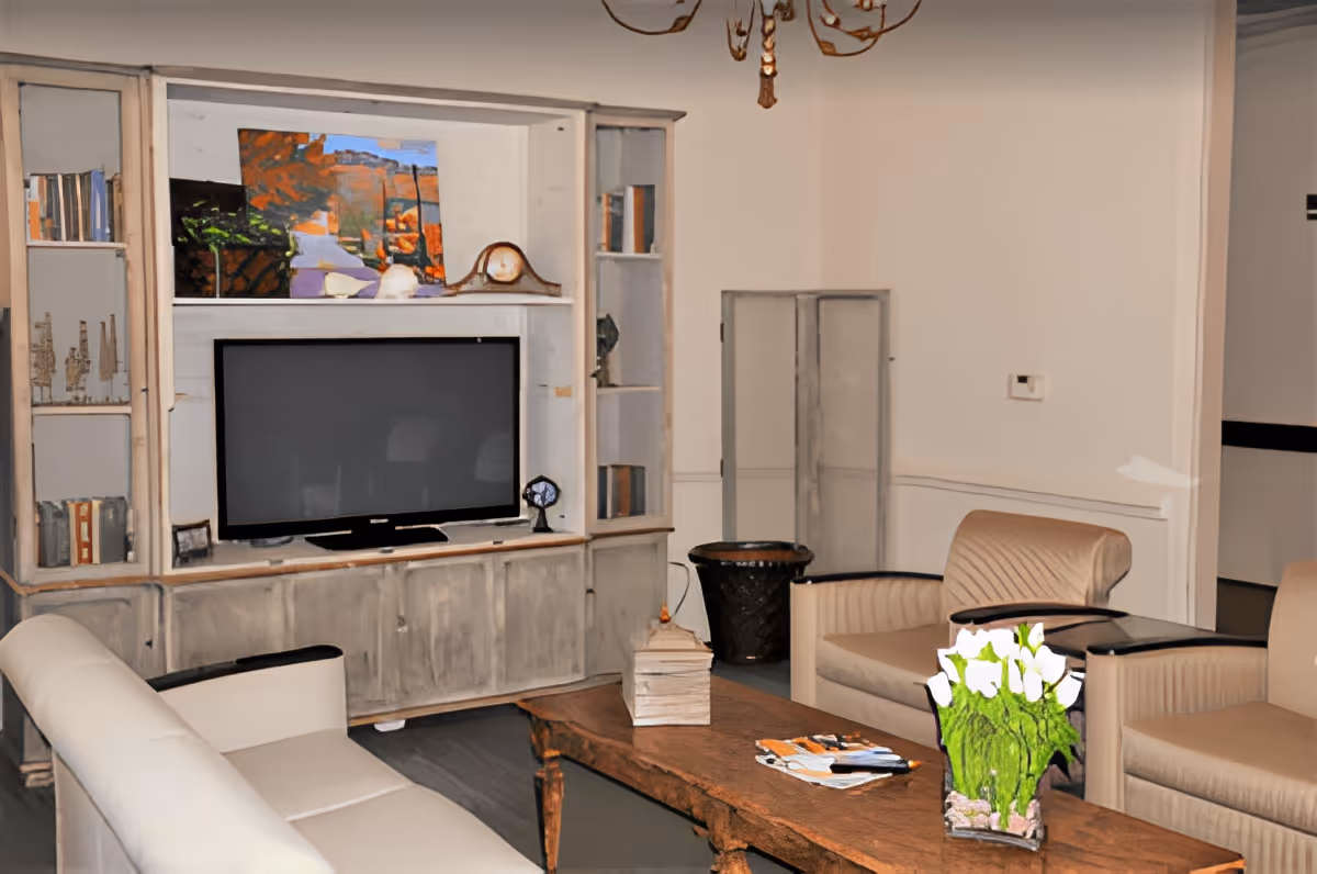 A cozy living room with a large flat-screen TV mounted on a rustic wooden entertainment center. The room features a white sofa, two beige armchairs, a wooden coffee table with a vase of white flowers and some magazines, and a decorative clock on the entertainment center. The walls are light-colored, and there is a chandelier hanging from the ceiling.