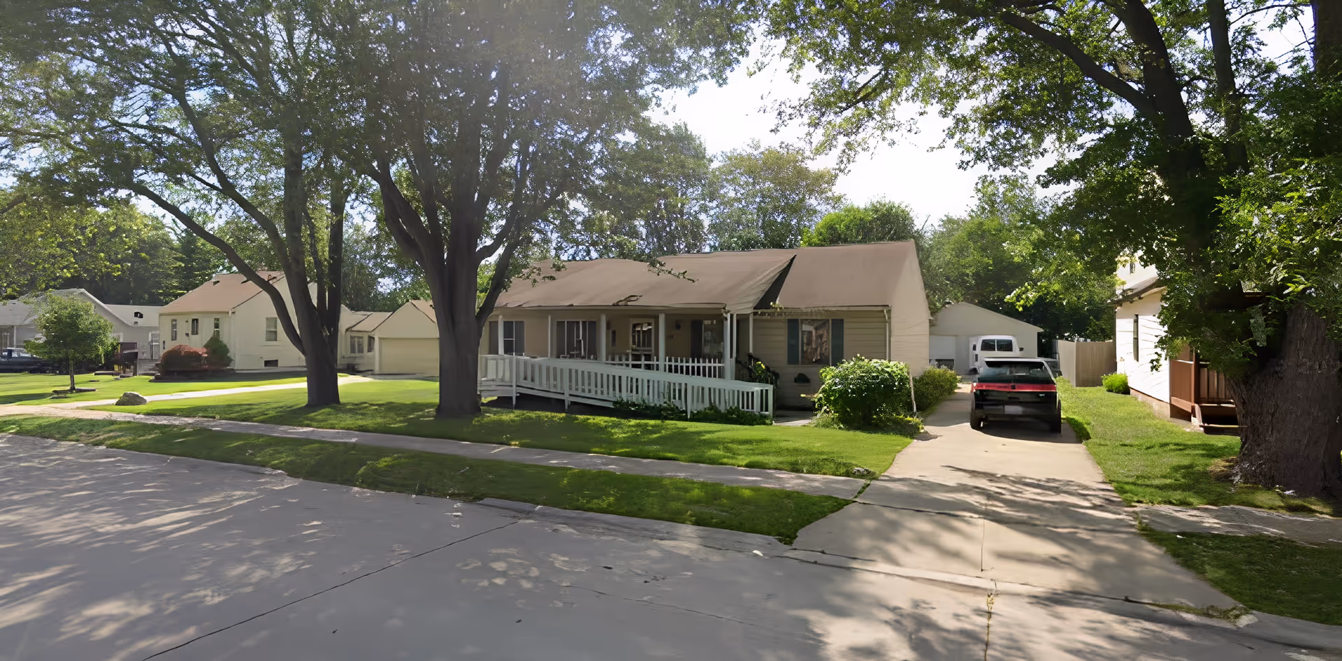 A single-story residential building with a front porch and a wheelchair ramp, surrounded by green grass and large trees. There is a driveway with a black vehicle parked on it, and neighboring houses are visible in the background under a partly cloudy sky.