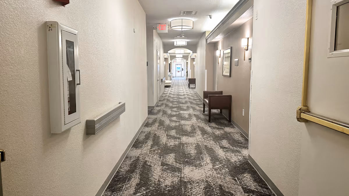 A long, well-lit hallway in a senior living facility with patterned carpet, beige walls, wall-mounted lights, and a few chairs along the right side. There is a fire extinguisher cabinet on the left wall and an exit sign visible at the far end of the corridor.